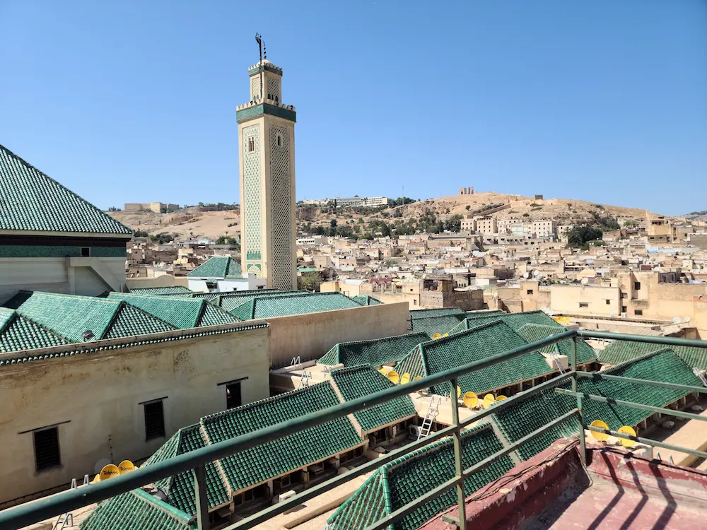 Fes medina from a rooftop: green-tiled roofs and the minaret of the Kairaouine mosque, with the old city and bare hills beyond under a clear blue sky
