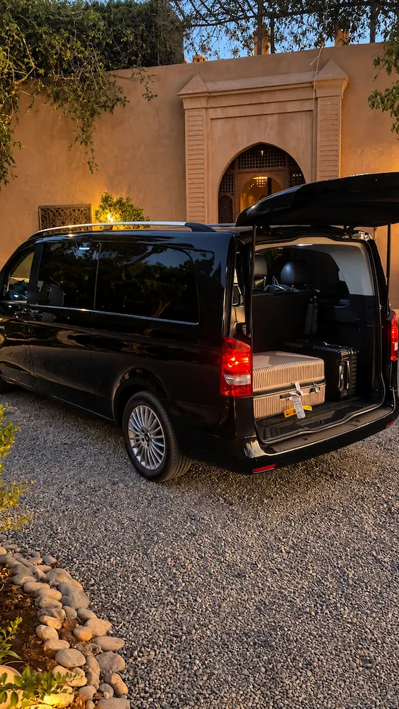 Black Mercedes van with the tailgate up and suitcases loaded in the boot, parked on gravel outside a riad gate at dusk before a Marrakech to Fes transfer
