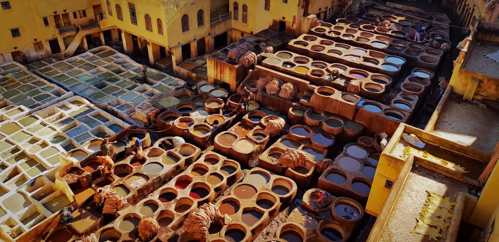 Traditional leather tanneries in the Fes medina seen from a terrace above: rows of round stone vats of dye and lime, workers between them, and hides drying on the roofs