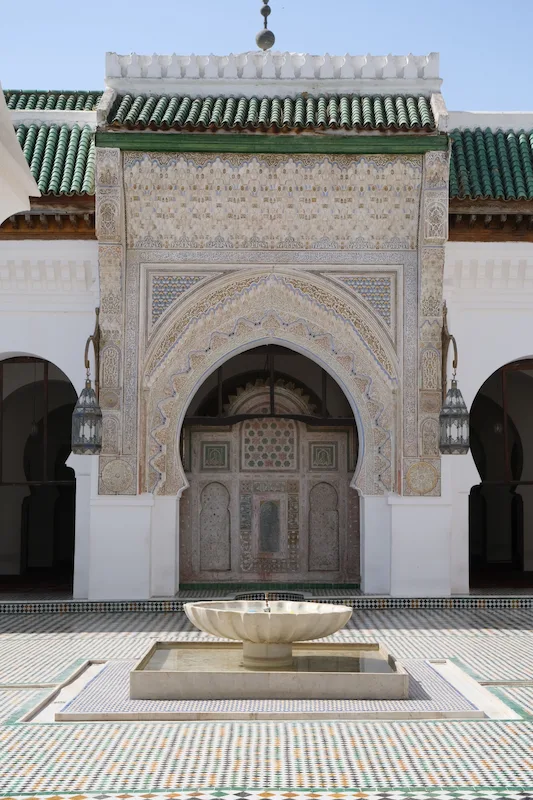 Courtyard of the Kairaouine mosque in Fes: a carved stucco arch under a green-tiled roof, hanging lanterns, and a marble fountain on a floor of zellige tiles