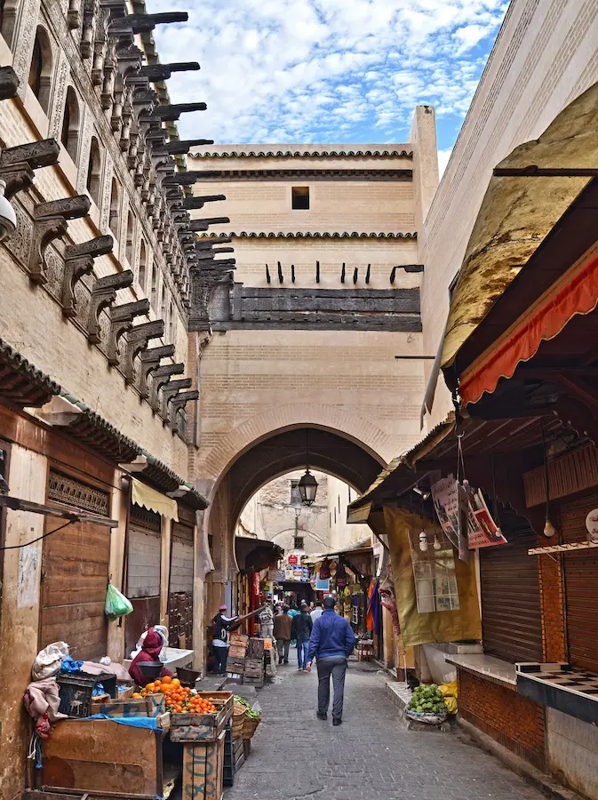 A market lane inside the Fès el-Bali medina, fruit stalls under a covered passage and an arched gateway ahead