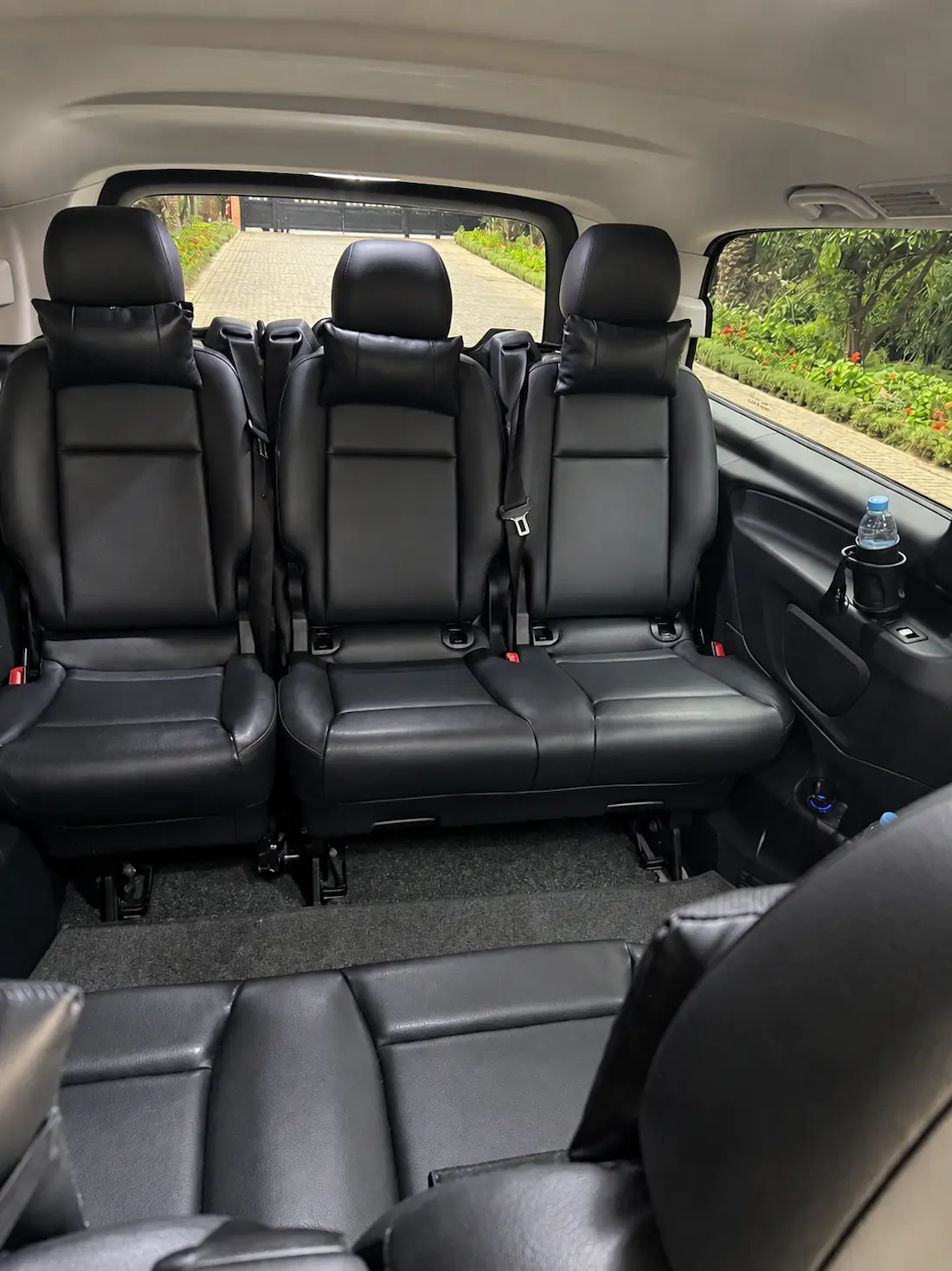 Rear cabin of the Mercedes van: three black leather seats with headrests and seatbelts, a water bottle in the door, and a garden drive through the back window