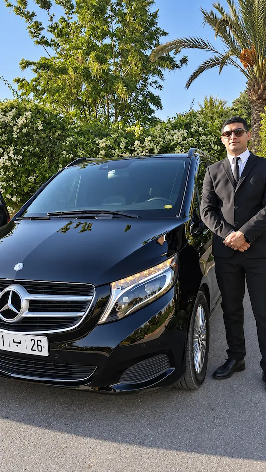 Licensed private driver in a dark suit beside a black Mercedes V-Class van under palm trees in Marrakech, the vehicle arranged for the Marrakech to Fes transfer